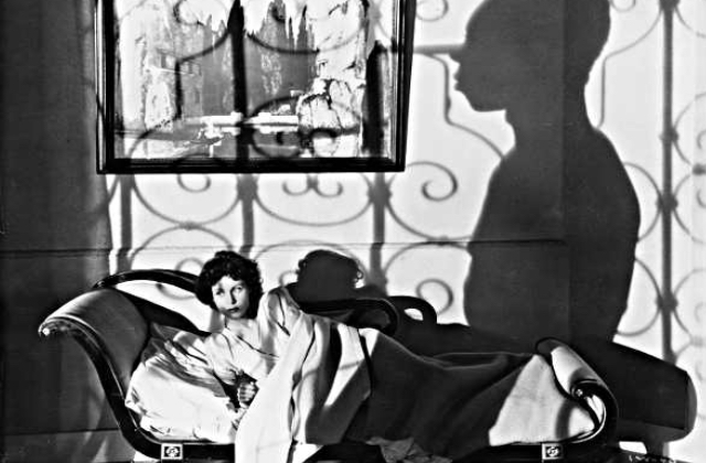 A black and white photo of a white woman lying on a divan, with the shadow of a black man on the wall behind her.